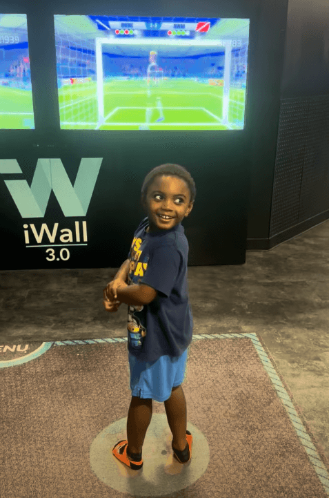 Smiling young boy looks back while standing before an interactive iWall soccer game screen.