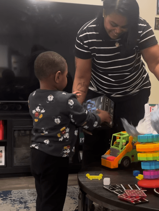 Smiling woman hands a boxed toy to a young boy wearing a Mickey Mouse sweatshirt.